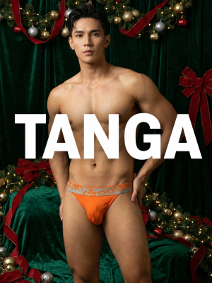Male model standing shirtless in orange tanga underwear against a green holiday backdrop with garlands and lights, large 'TANGA' text across the image.